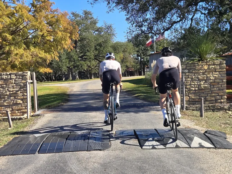 Two riders crossing a cattle guard at a ranch gate with Texas and US flags.